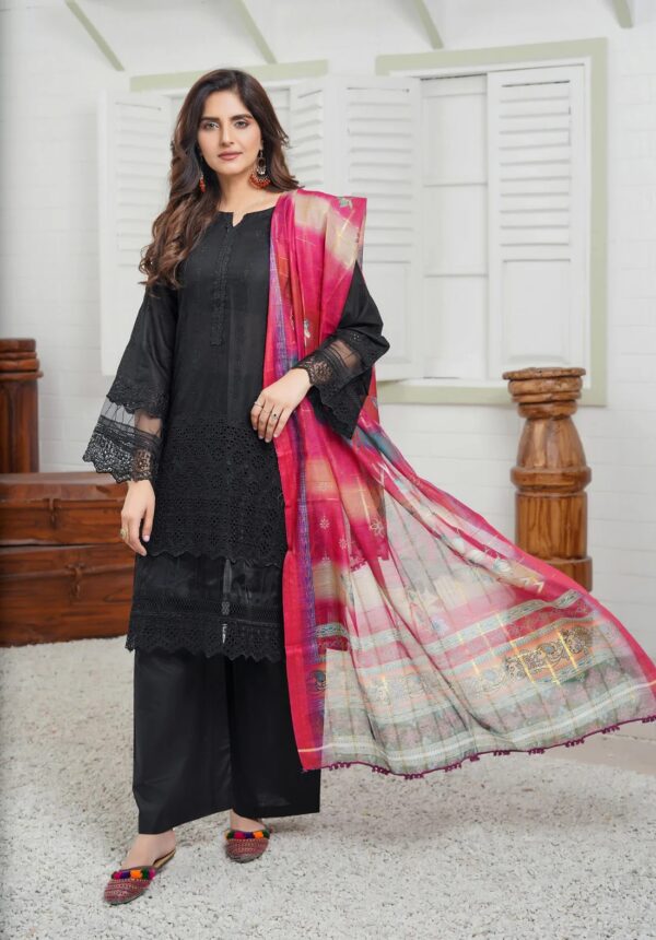 A woman in a black suit in a multi-color dupatta stands in a room in front of a window, posing for a photo shoot from the front of the black suit.